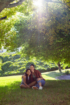 African American mother and daughter sitting and hugging on grassy lawn under shade tree smiling