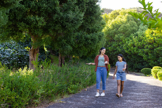 African mother and adolescent daughter walking along paved garden path past tree trunk, copy space