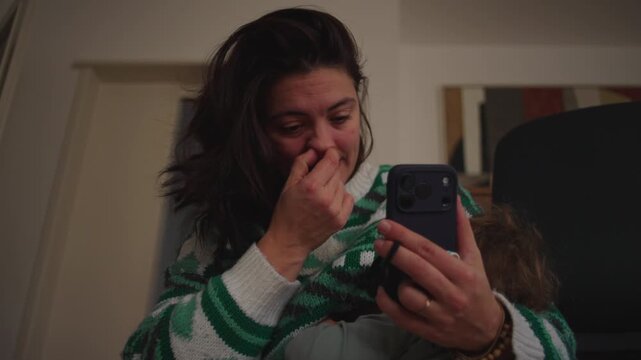 Mother on smartphone video call while breastfeeding toddler and scratching nose, candid home parenting moment showing multitasking and everyday reality