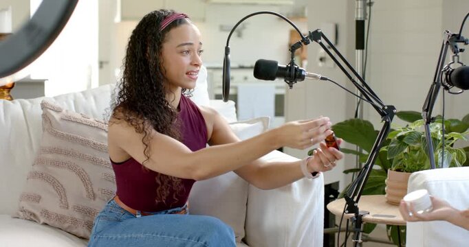 African American women on sofa, host offering dropper jar for tester smelling and rubbing near mic