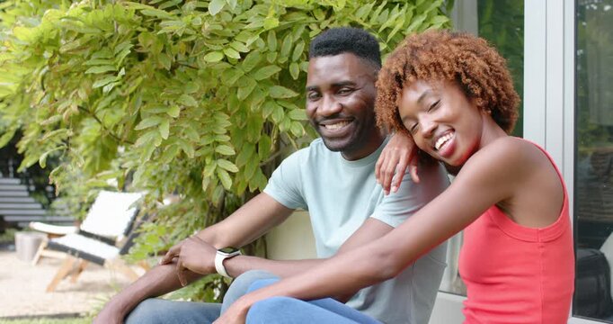 Diverse couple posing on porch bench, woman reaching up, resting head on man's shoulder, smartwatch