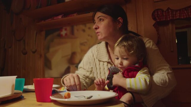 Mother speaking to someone off camera while holding toddler during dinner at wooden table inside cozy alpine chalet home