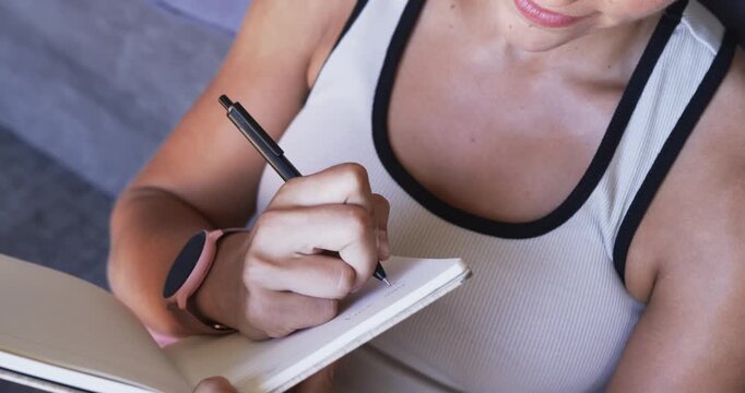 Woman steadying notebook while writing notes with black pen on sofa, wearing pink smartwatch