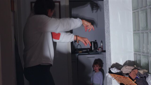 Playful mother forming animal shadow figures with hands on wall while toddler watches in kitchen, intimate family moment of creativity and affection