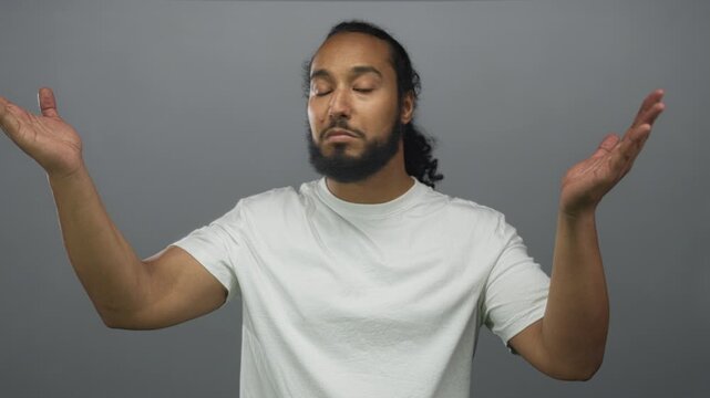 Bearded man shrugging with raised palms and closed eyes in a gray studio, neutral expression and relaxed posture; indifference.