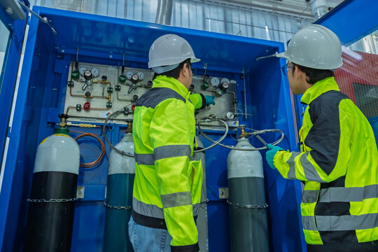 Two Asian engineers inspecting industrial gas manifold system. Professional technicians checking pressure gauges and monitoring oxygen nitrogen argon cylinders at manufacturing plant.