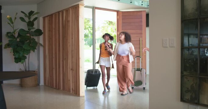 Diverse female friends entering pivot door, pointing up, pulling suitcases to inspect home