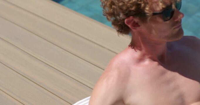 Man reclining on towel on wooden deck by pool, camera pulling back, propping up, wearing sunglasses