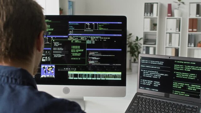 Over shoulder shot of young adult male IT programmer reviewing code on multiple computer screens solving problems with software while working at desk in tech company office