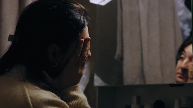 Woman rubbing tired face while looking at reflection in bathroom mirror, late evening fatigue and quiet anxiety about aging after long day