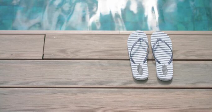 Camera is panning down to reveal striped flip-flops sitting on wood deck beside rippling aqua pool