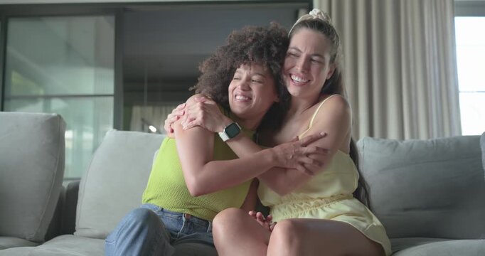 Diverse female friends sitting on gray sofa at home, talking leading to hugging, showing affection