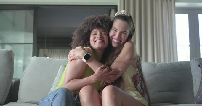 Diverse female friends laughing on sofa at home right woman touching scrunchie hugging showing love