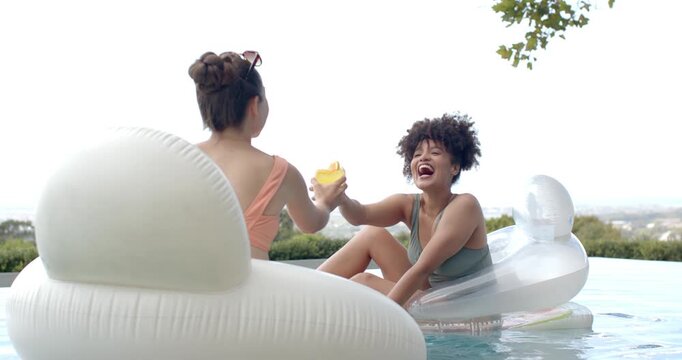Diverse female friends in swimwear after laughter clinking yellow cups on floats at infinity pool