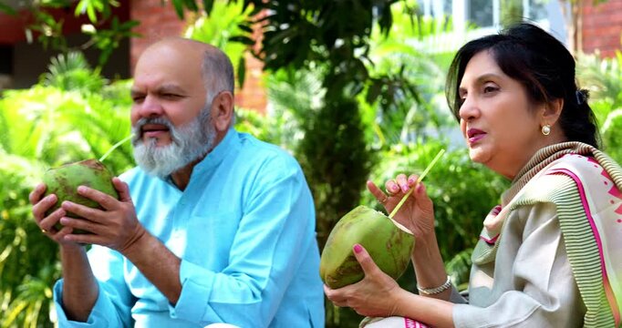 Indian senior couple having fresh coconut water together in nature sitting in home garden, drinking with straw from green nariyal and enjoying peaceful outdoor conversation