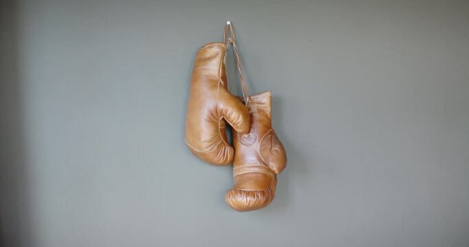 Tan boxing gloves are gently swaying from slight draft on hook on gray wall, laces shifting