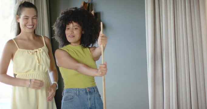 Two women standing at home holding wooden pole, spotting outside and pointing at window curtains