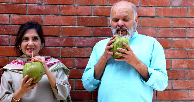 Indian senior couple having fresh coconut water together in nature sitting in home garden, drinking with straw from green nariyal and enjoying peaceful outdoor conversation