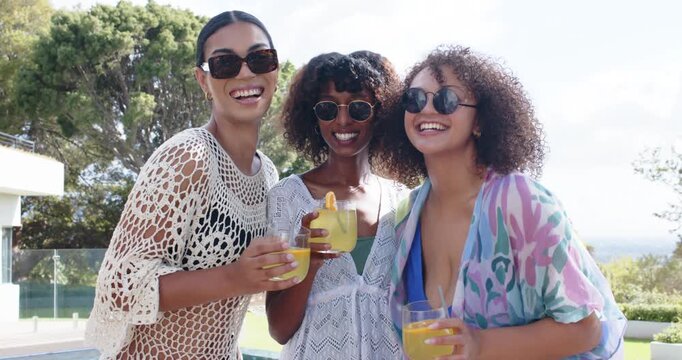 3 African American women prompted by camera raising orange drinks at poolside in wraps sharing joke
