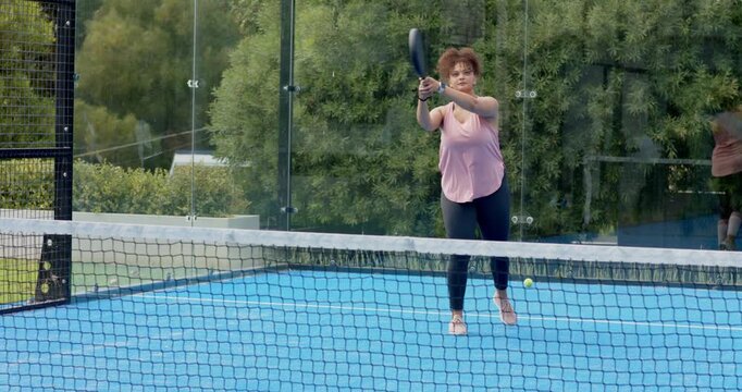 African American woman in pink top reacting to bounce, returning with padel racket on glass court