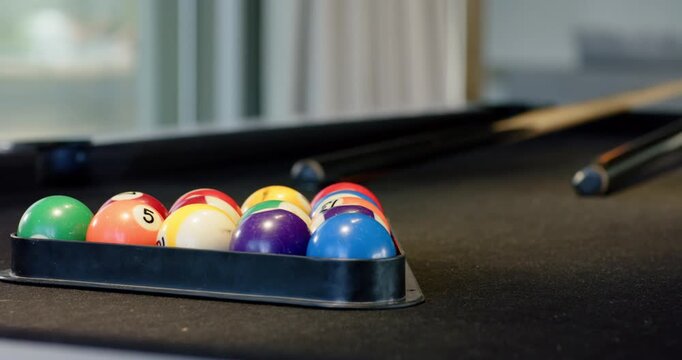 Camera is pulling back revealing racked billiard balls on black felt table near window showing cues