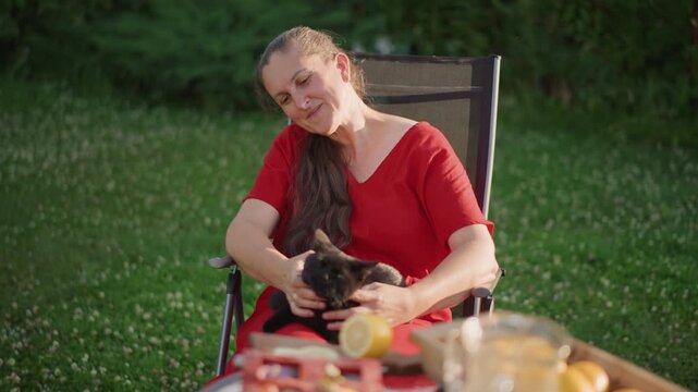 Caucasian woman in red dress smiling while cradling black cat at backyard picnic table, colorful snacks and drinks in foreground, warm afternoon light, playful pet interaction, hospitality vibe,
