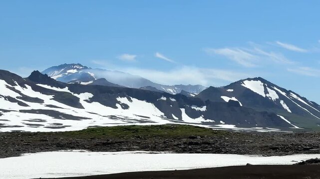 Mutnovsky volcano in Kamchatka rising above dark volcanic ground and summer snowfields under a clear blue sky, captured in bright daylight in a remote highland setting. Suitable for travel, wilderness