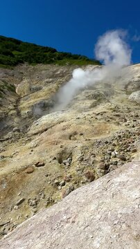 Active geothermal hillside with white steam vents, mineral-stained ground, and rocky terrain in Kamchatka, Russia, captured on a clear sunny day. Suitable for travel, volcanic activity, geothermal ene