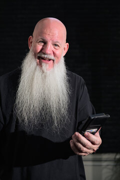 Portrait of a happy bald man with a long white beard using phone against a dark background
