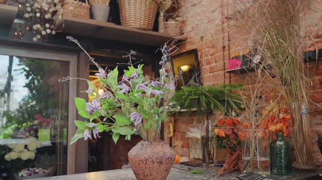 Cozy florist studio with rustic brick and handcrafted vase of purple clematis blooms on wooden counter, soft ambient light, shelves of dried botanicals and pottery, intimate closeups of petals, stems