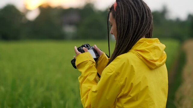 Young photographer with dreadlocks, wearing a vibrant yellow raincoat, capturing stunning images in a scenic field at sunset, using a professional camera to document the beauty of nature