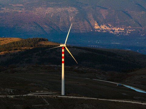 Aerial view of a striking wind turbine with red and white stripes piercing the skyline against a backdrop of rolling hills and distant mountains, Cocullo, Abruzzo, Italy.