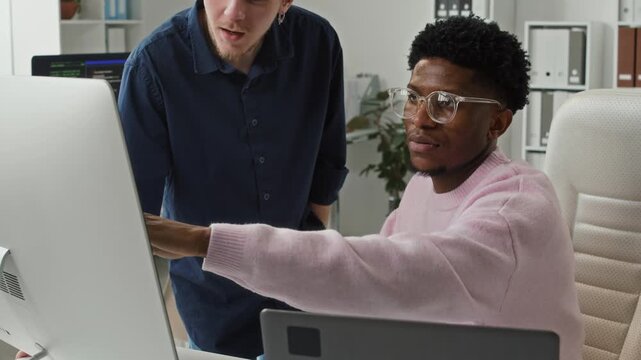 Focused young Black male programmer with glasses pointing at computer screen reviewing code for bugs while brainstorming ideas with peer colleague at workstation in shared space office