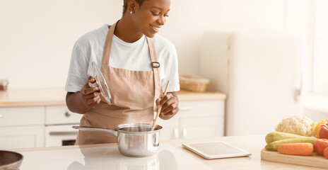 A person stands in a bright kitchen cooking vegetables in a pan. The individual uses a tablet on the counter to follow a recipe while mixing ingredients. Fresh produce is displayed. © Prostock-studio