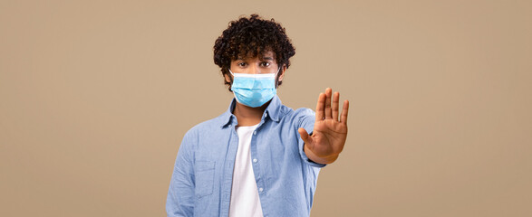 A young man stands in a studio against a bright yellow background. He has curly hair and wears a mask. He raises his hand as a gesture, looking directly at the viewer. © Prostock-studio