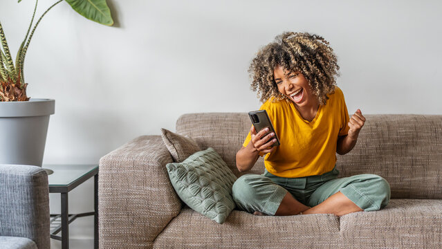 Excited happy young black woman holding smart phone device sitting on sofa at home - Happy satisfied female looking at mobile smartphone screen gesturing yes with clenched fist - Technology concept