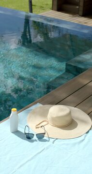 Vertical video: Light breeze rippling pool as hat, sunglasses, sunscreen are resting on towel