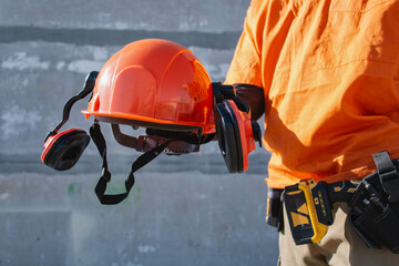Construction worker holding orange safety helmet with ear protection at industrial site. Workplace safety and occupational protection concept suitable for engineering services and building.