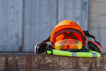 Orange hard hat with protective goggles, earmuffs and high visibility vest against industrial background. Personal protective equipment for construction workers and industrial safety.