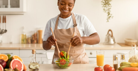 A woman stands in a kitchen mixing a fresh salad. She uses wooden chopsticks and smiles while surrounded by vegetables, drinks, and other ingredients on the counter. © Prostock-studio