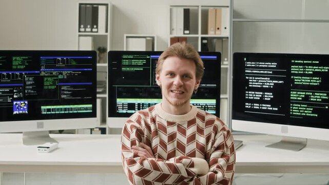 Portrait of young adult male IT developer in casual clothes turning to camera with smile while sitting against coding setup in modern office