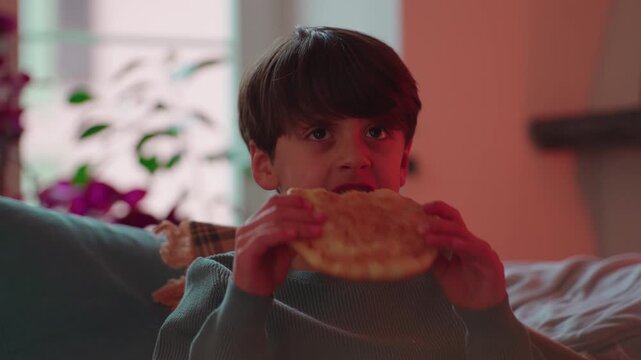 Young boy sitting on couch eating slice of pizza with both hands, relaxed evening at home, candid childhood moment of comfort food and quiet enjoyment