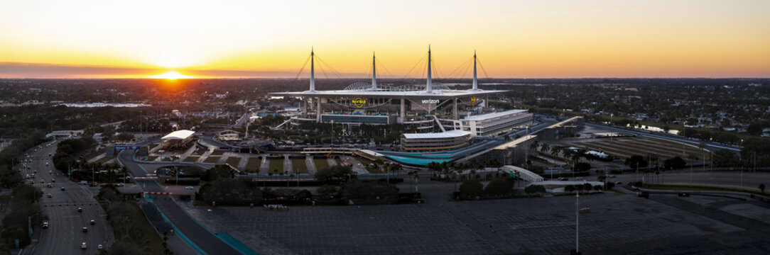 Miami Gardens, USA - 23 February 2026: Aerial view of Hard Rock Stadium illuminated by the setting sun, casting long shadows across the vast parking lot and cityscape.