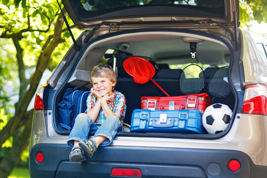 Adorable little kid boy sitting in car trunk just before leaving for summer vacation with his parents. Happy child with suitcases and toys going on journey, real life, real people