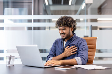 Fototapeta premium A young Indian man, twisted in pain, sits in an office at a desk with a laptop and holds his hand to his chest