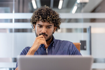 Thoughtful and focused young Indian man working in the office on a laptop
