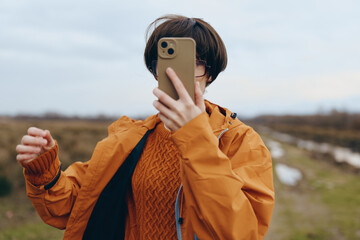 Young woman lifestyle taking selfie with smartphone outdoors in casual orange jacket and sweater....