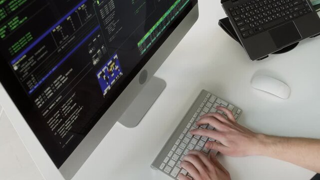 High angle shot of unseen young adult male IT specialist typing on computer keyboard editing code displayed on multiple screens while programming at workstation in office