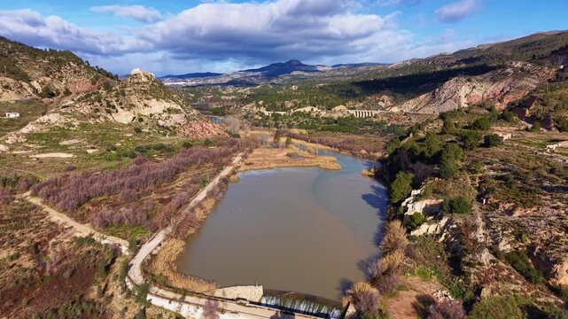 Aerial view of the historic castle of Domeno overlooking a winding river valley in eastern Spain, with rugged hills, forest patches, and a railway viaduct crossing the Mediterranean landscape