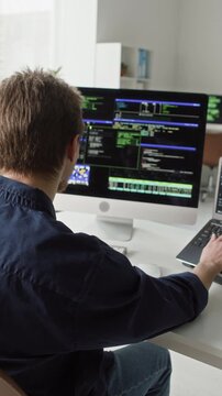 Vertical shot of focused young adult male IT developer in glasses typing on laptop and desktop computer at programming desk writing software code in modern office
